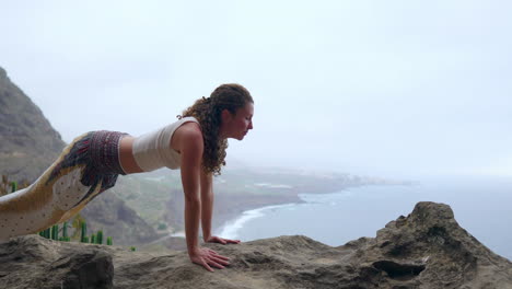 Positioned-on-a-cliff's-edge,-the-woman-practices-a-dog-pose,-overlooking-the-ocean,-as-she-breathes-in-the-sea-air-during-her-yoga-journey-across-the-islands